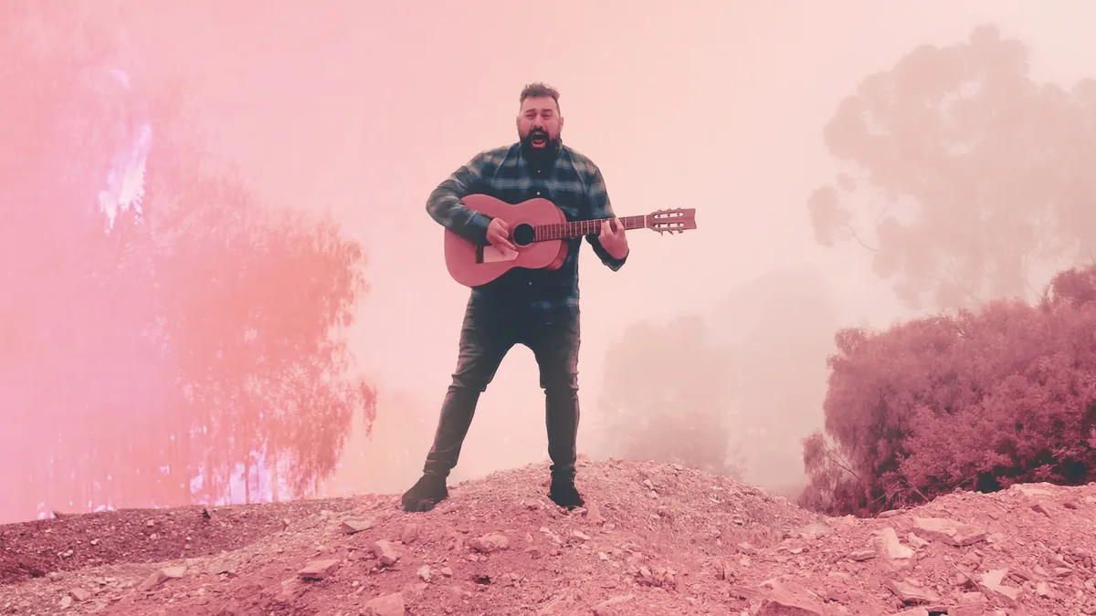 Ceschi, a bearded man, stands on a dirt mound playing an acoustic guitar against a hazy, pink-tinged background.