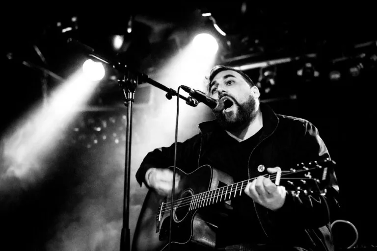 Black and white photo of Ceschi playing an acoustic guitar and singing into a microphone on a dimly lit stage.