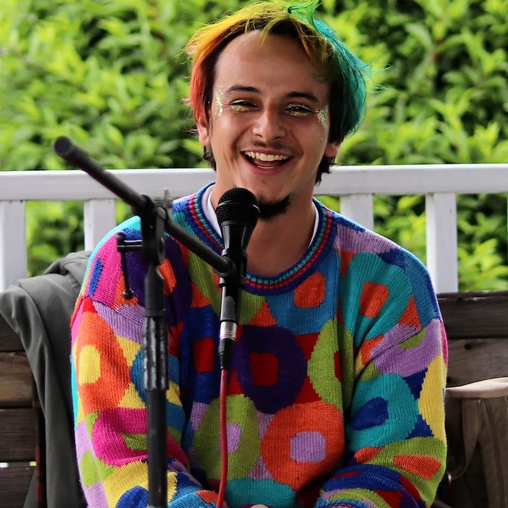 Myles Bullen with rainbow hair and glitter eyelids, wearing a colorful sweater, smiling into a microphone.