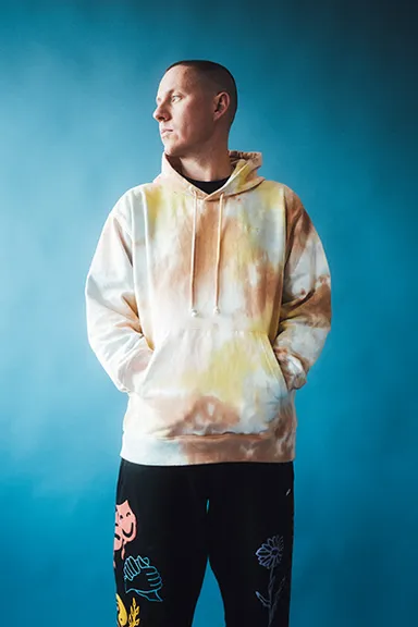 Factor Chandelier in a tie-dye hoodie, looking to the right against a blue background.