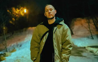 Factor Chandelier stands outdoors at night, wearing a beige jacket over a black hoodie, looking directly at the camera.