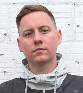 Headshot of Factor Chandelier, a man with short brown hair, wearing a grey and black hoodie, against a white brick wall.